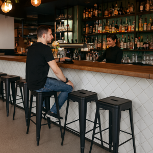 Super-Seat | Zwarte metalen barkruk, man in zwart, houten bar, flessenrek achter vrouw achter de bar. | Horecameubilair