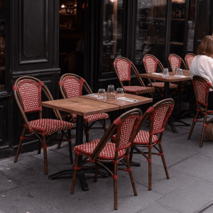 Super-Seat |Terrasstoelen en -tafelsRode rieten terrasstoelen, bistrotafels met glazen/bestek; 1 persoon zit rechts alleen. | Horecameubilair
