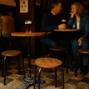 Super-Seat | Wat je gaat tegenkomen zijn stoelen, tafels, barkrukken, picnick tafels en andere soort tafels en stoelen. | Houten tafel met twee bruine Vanitas schoolkrukken op dambordvloer; man en vrouw drinken bij warme verlichting in pub. | Horecameubilair