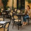 Super-Seat | Een vrouw zit alleen aan een cafétafel op een Chopin Noir Thonet (terras) stoel met houten frame, metalen terrastafels, glas water; gele muren, groene planten. | Horecameubilair
