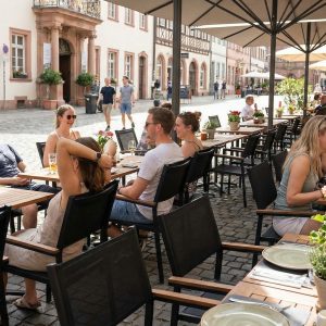 Super-Seat | Terrasstoelen Sunny Zwart | Zwarte textileen stoelen met armleun, metalen tafels, houten plantenbakken en parasols op straat. | Horecameubilair