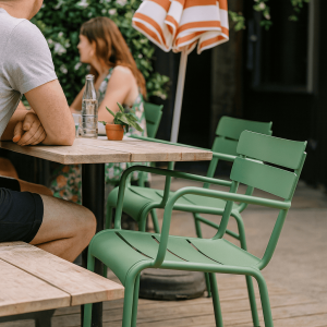 Super-Seat |Houten tafel, groene metalen armstoelen, oranje-wit gestreepte parasol, fles en plant op tafel. | Horecameubilair