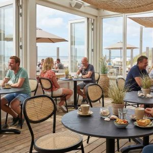 Super-Seat | Zwarte stoelen en barkrukken rond zwarte HPL roest/koper tafels, uitzicht op duinen en zee. | Horecameubilair