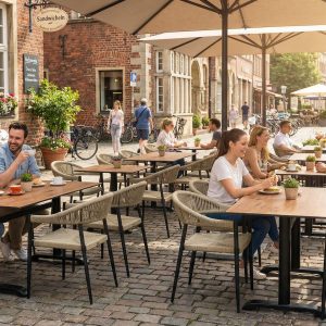 Super-Seat | Je ziet terrastafels van teakhout, metalen stoelen, bloemenbakken en grote parasols op een klinkerstraat. | Horecameubilair