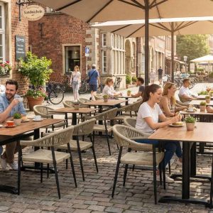Super-Seat | Je ziet terrastafels van teakhout, metalen stoelen, bloemenbakken en grote parasols op een klinkerstraat. | Horecameubilair