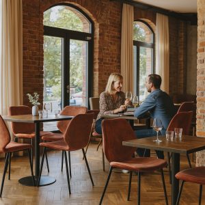 Super-Seat |Een vrouw en een man zitten aan een restauranttafel... Zwarte vintage leren stoelen & rode stoelen bij tafels, bakstenen muur, grote ramen. | Horecameubilair