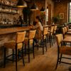 Super-Seat | Een vrouw zit alleen aan een houten bar in een warm verlicht, bakstenen restaurant met een drankje in haar hand. | Okergele velours Lisa Rome barkrukken met zwarte metalen poten aan een houten bar, warme sfeer en bakstenen muur. | Horecameubilair