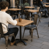 Super-Seat | Horeca Terras Tafels Tivoli Teak 70x70cm | Persoon in witte trui zit alleen aan teakhouten tafel met metalen onderstel, andere tafels verspreid. | Horecameubilair