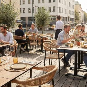 Super-Seat | HPL Horeca Tafelbladen Kasteel Eiken 80x80cm Houten eikenkleurige tafels met stalen frames en stoelen, omringd door planten en lampjes. | Horecameubilair