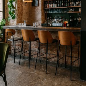 Super-Seat | Een bar met een stenen vloer, bakstenen muur en vier New Yorkse design barkrukken hout zwart leer. | Vier zwarte leren barkrukken met houten poten; bar met stenen vloer en binnenmuur. | Horecameubilair